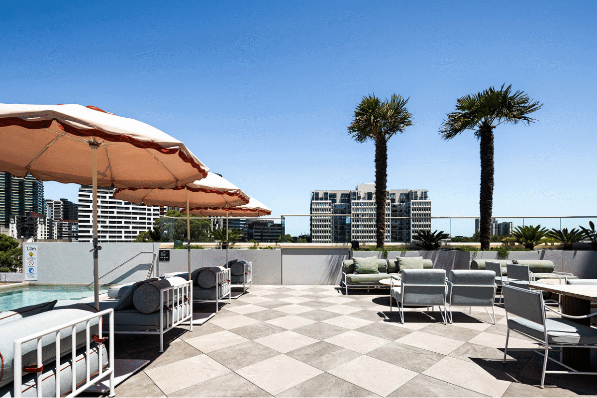 Modern rooftop terrace with checkered stone flooring, soft grey outdoor seating and blush umbrellas under a clear blue Sydney sky – styled with a contemporary coastal aesthetic by BOWERBIRD Interiors.