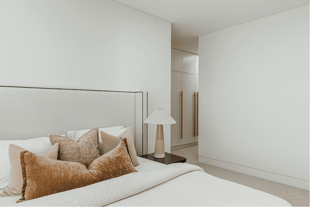 Minimal bedroom styled with a tall upholstered bedhead, layered neutral cushions, a textured throw, and a bedside lamp, with built-in wardrobes in the background.