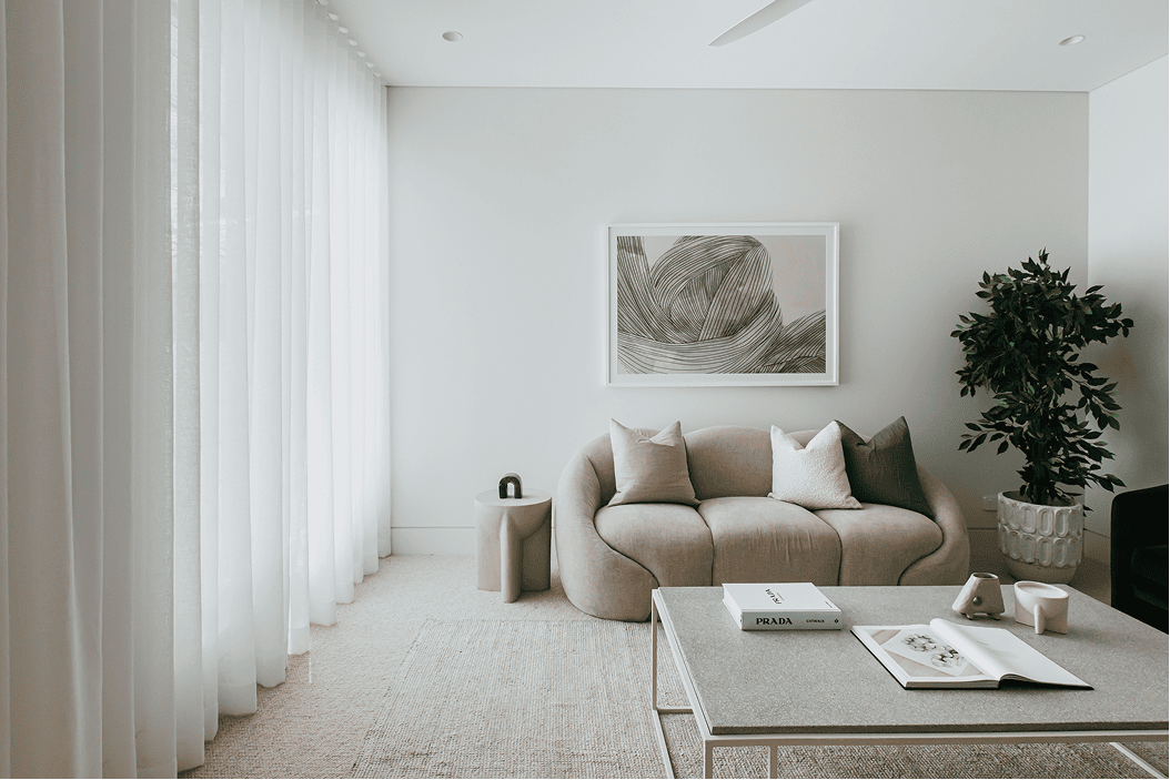 Soft, neutral living room with sheer floor-to-ceiling curtains, a curved beige sofa with layered cushions, framed artwork, a potted tree, and a coffee table styled with books and ceramics.