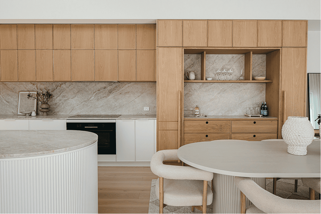 Light-filled kitchen and dining area with pale timber cabinetry, stone splashback, a white fluted island bench, built-in bar niche, and a round dining table with upholstered chairs.