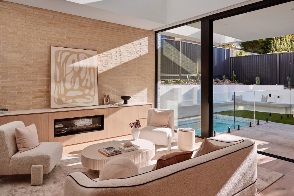 A cozy and stylish living room with plush, modern furniture. The soft, neutral tones of the seating area are complemented by a brick wall and a minimalist, low-profile fireplace. Glass doors open to an outdoor area with a pool, framed by lush greenery and low fencing.