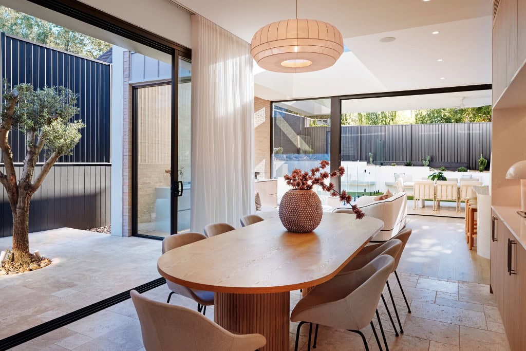 A dining area with an elegant wooden table surrounded by soft, neutral chairs. The space is open to a courtyard garden, with large glass doors leading to an outdoor seating area, framed by sheer white curtains. A round pendant light hangs above the table.