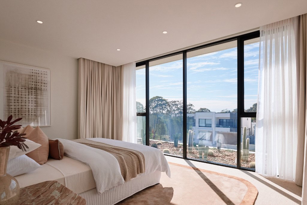 A spacious and tranquil bedroom with a neutral color scheme. A large bed with soft, layered bedding is framed by floor-to-ceiling windows that offer a view of a landscaped garden. The space is bathed in natural light, creating a calm and airy atmosphere.
