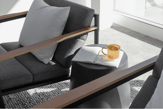 Sunlit commercial lounge corner with a charcoal armchair, pale grey cushion, and a small black side table holding an open notebook and amber glass mug.
