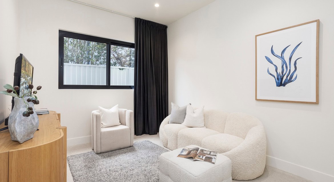 A cozy, minimalist living area featuring soft, cream-colored furniture, including a curved couch and a single armchair with plush cushions. A small, light gray rug covers the floor, and a decorative magazine is placed on a matching ottoman. The room has a black-framed window with a view of greenery, and a dark curtain hangs next to it. A framed watercolor print of blue leaves is displayed on the wall, adding a touch of color to the neutral tones.