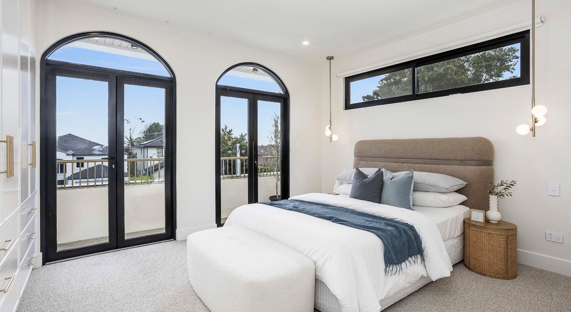 A stylish bedroom featuring a plush bed with a beige headboard and blue throw, accented by soft gray pillows. The room has large, black-framed windows and French doors with a balcony view. A modern light fixture hangs above the bed, and a woven rattan nightstand sits beside it, holding decorative items. The room has a bright, airy atmosphere with neutral tones and carpeted flooring.