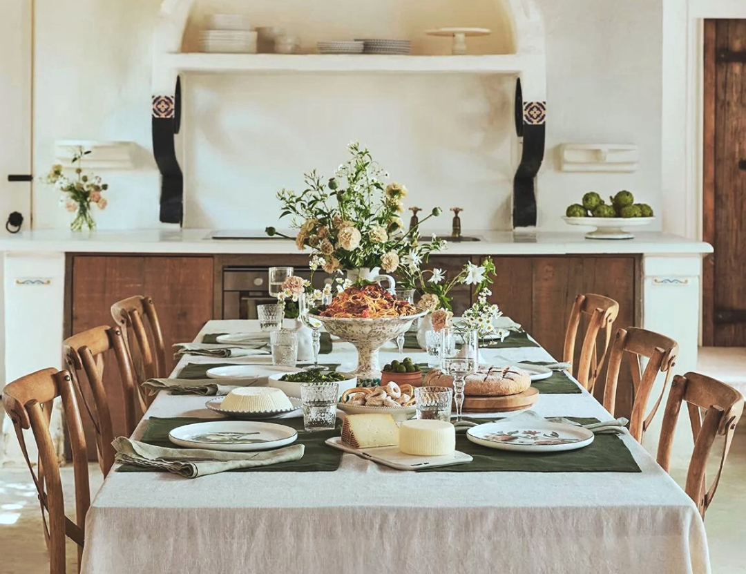 A Rustic Dining Room Set for a Meal, Featuring a Long Table With a Linen Cloth, Wooden Chairs, and a Plaster Archway in the Background